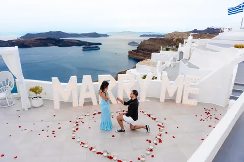 Surprise marriage proposal on a Santorini caldera terrace with MARRY ME letters and a rose petal heart